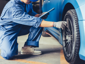A man inspects the tire of a car with gloves and a clipboard.